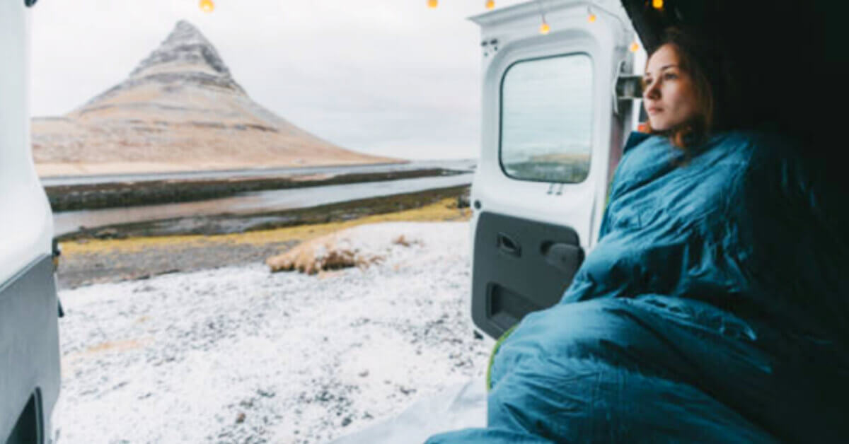 Women sitting in a campervan with the doors open in the snow snuggled up in a sleeping bag 